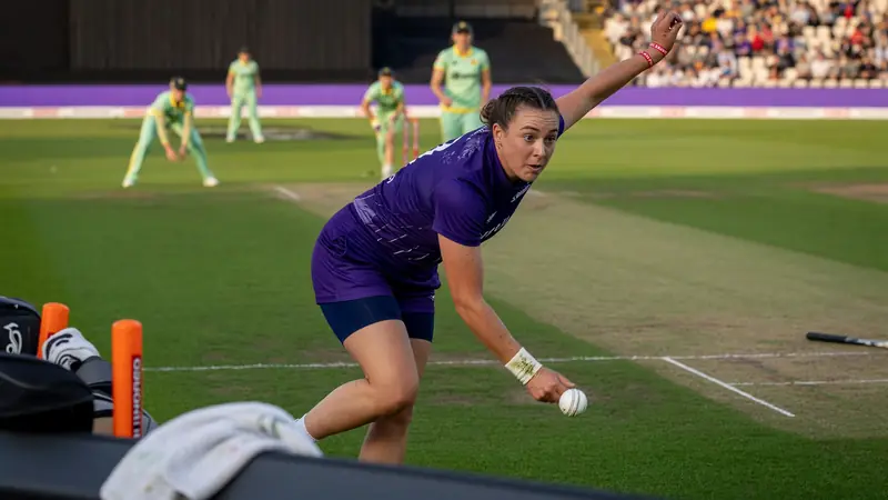 Women's cricket batter driving through cover under floodlights with the wicketkeeper standing up and the slip cordon watching